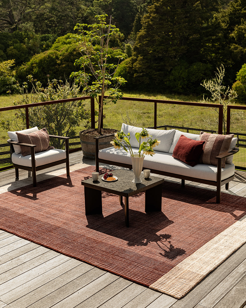 Baya Bedrock Indoor and Outdoor Floor Rug in Claret, full-width view on a deck beneath a sofa, armchair and coffee table, showing the rug's rust-red field and pale woven border.