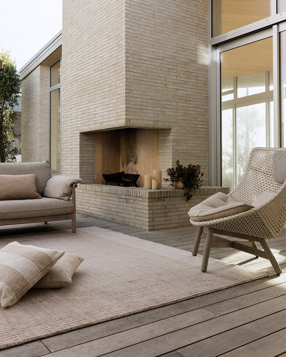 Baya Bedrock Indoor and Outdoor Floor Rug in Sand, styled on an outdoor deck beside a brick fireplace with a woven rattan armchair, linen sofa and floor cushions.
