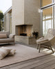 Baya Bedrock Indoor and Outdoor Floor Rug in Sand, styled on an outdoor deck beside a brick fireplace with a woven rattan armchair, linen sofa and floor cushions.