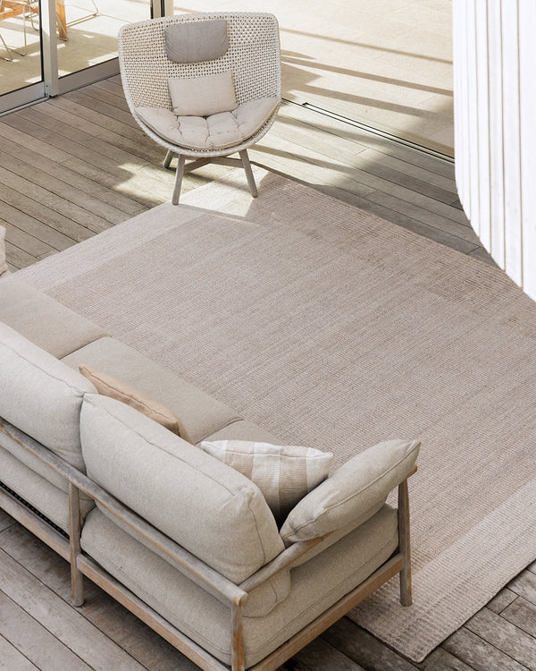 Baya Bedrock Indoor and Outdoor Floor Rug in Sand, overhead view on a covered timber deck beneath a woven rattan chair and linen sofa, showing the rug's pale sand field and lighter striped border.