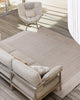 Baya Bedrock Indoor and Outdoor Floor Rug in Sand, overhead view on a covered timber deck beneath a woven rattan chair and linen sofa, showing the rug's pale sand field and lighter striped border.