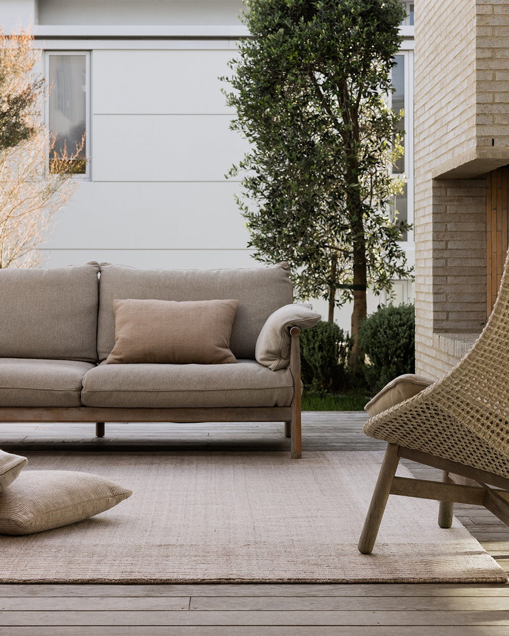 Baya Bedrock Indoor and Outdoor Floor Rug in Sand, styled on an outdoor deck beneath a linen sofa and woven rattan armchair, with floor cushions and an olive tree against a brick and weatherboard home.
