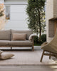 Baya Bedrock Indoor and Outdoor Floor Rug in Sand, styled on an outdoor deck beneath a linen sofa and woven rattan armchair, with floor cushions and an olive tree against a brick and weatherboard home.