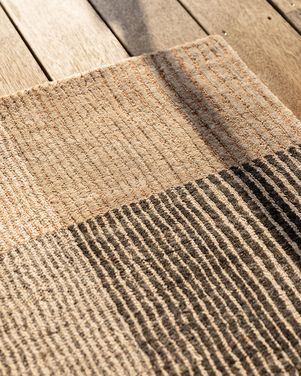 Baya Bedrock Indoor and Outdoor Floor Rug in Zinc, sunlit corner detail on an outdoor timber deck showing the pale sand striped border meeting the charcoal-grey striped field.