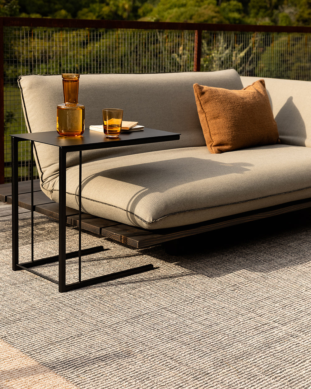 Baya Bedrock Indoor and Outdoor Floor Rug in Zinc, closer view of the outdoor daybed setting with a decanter and glass on the black side table, showing the rug's charcoal-flecked texture and sand-toned border edge.