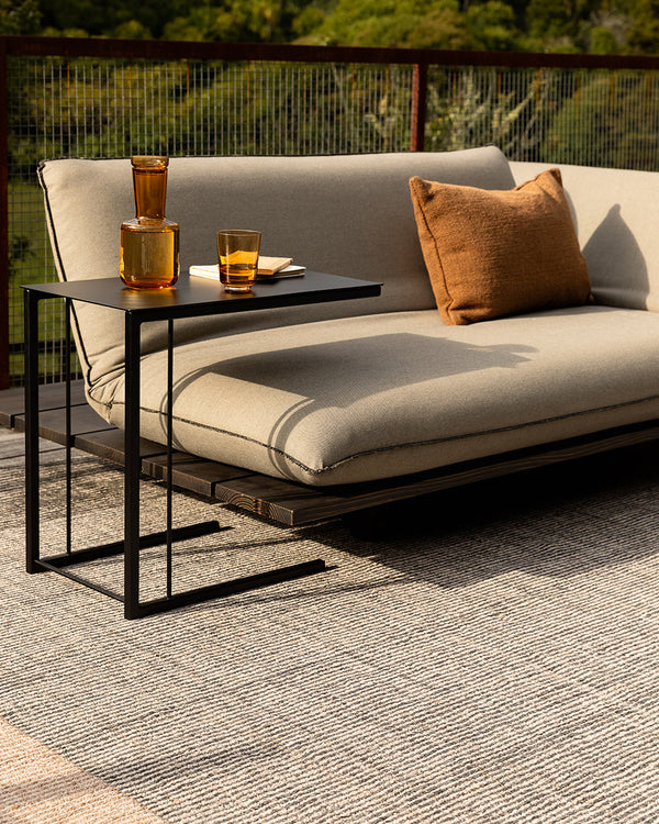 Baya Bedrock Indoor and Outdoor Floor Rug in Zinc, closer view of the outdoor daybed setting with a decanter and glass on the black side table, showing the rug's charcoal-flecked texture and sand-toned border edge.