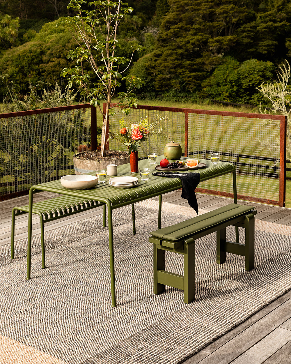 Baya Bedrock Indoor and Outdoor Floor Rug in Zinc, styled beneath an olive-green outdoor dining table and benches set for a meal, with a potted olive tree and flowers on a timber deck.