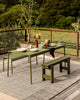 Baya Bedrock Indoor and Outdoor Floor Rug in Zinc, styled beneath an olive-green outdoor dining table and benches set for a meal, with a potted olive tree and flowers on a timber deck.
