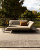 Baya Bedrock Indoor and Outdoor Floor Rug in Zinc, styled on an outdoor deck at golden hour beneath a low daybed-style sofa with a mustard cushion and black side table, with trees and a garden fence behind.