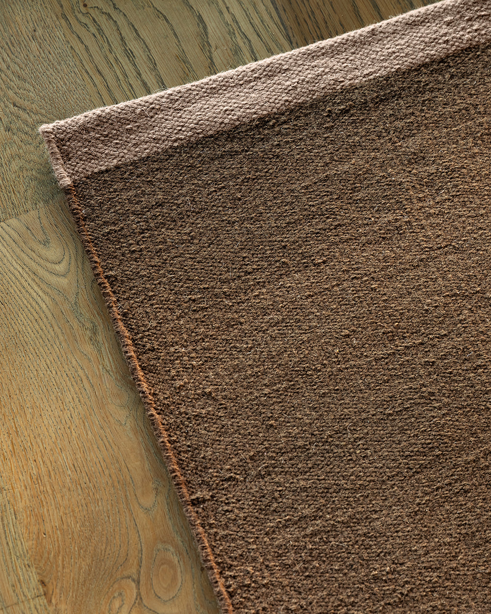 Baya Carla NZ wool floor mat in Cocoa, folded corner showing the weave and underside on a wood floor.