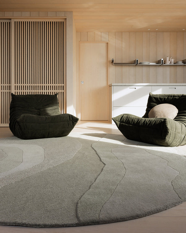 Baya Edgewater NZ wool floor rug in Pistachio, round, styled in a timber-panelled room with two dark green lounge chairs.