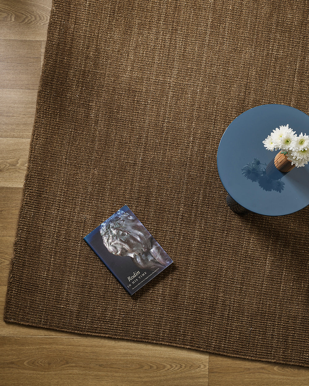Overhead styling shot of the Baya Linden Floor Rug in Ironbark beside a round side table with a book and a vase of white chrysanthemums.