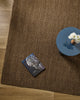 Overhead styling shot of the Baya Linden Floor Rug in Ironbark beside a round side table with a book and a vase of white chrysanthemums.
