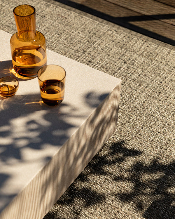 Baya Ormond outdoor rug in Mineral, styled on a deck under a pale stone table with an amber glass carafe and tumblers.