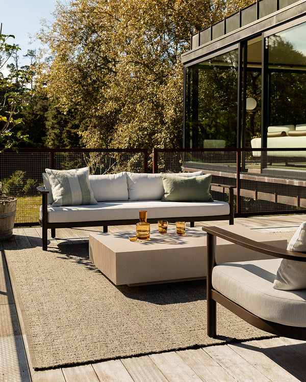 Baya Ormond outdoor rug in Mineral, styled on a timber deck beneath an outdoor sofa and coffee table, with a garden behind.