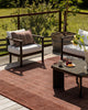 Baya Shore outdoor lumbar cushion in Claret styled on an outdoor armchair, with a rust-red rug and potted tree on the deck