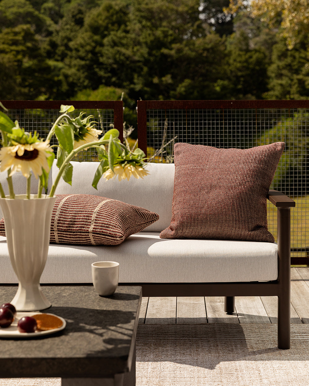 Baya Shore outdoor lumbar cushion in Claret styled on an outdoor sofa beside a second, plain textured cushion, with sunflowers and a fruit plate