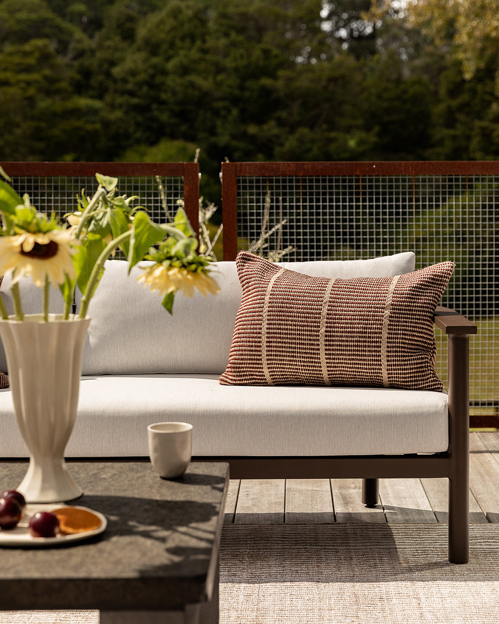 Baya Shore outdoor lumbar cushion in Claret styled alone on an outdoor sofa, with sunflowers and a fruit plate