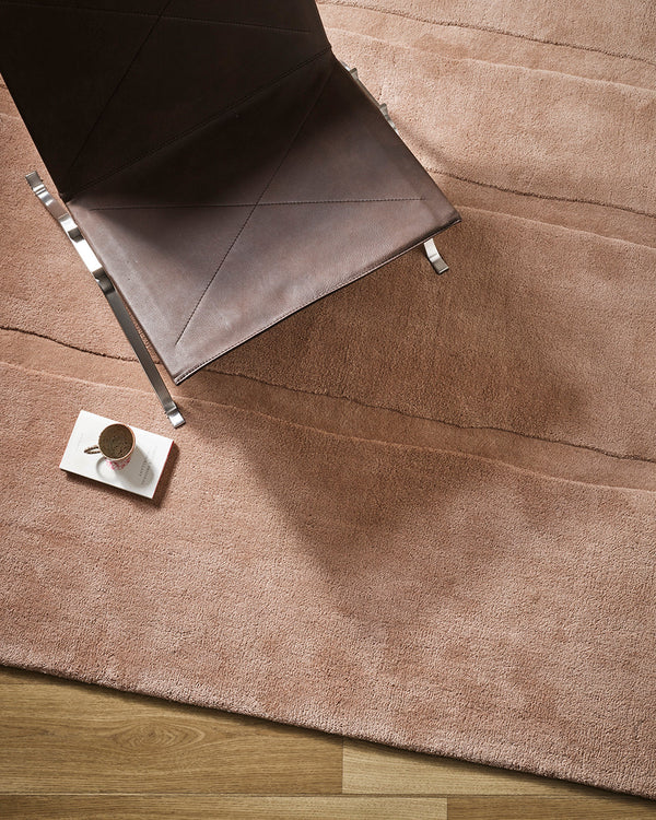 Baya Strata NZ wool floor rug in Clay, overhead view beneath a leather chair with a coffee cup on a saucer, on a timber floor.