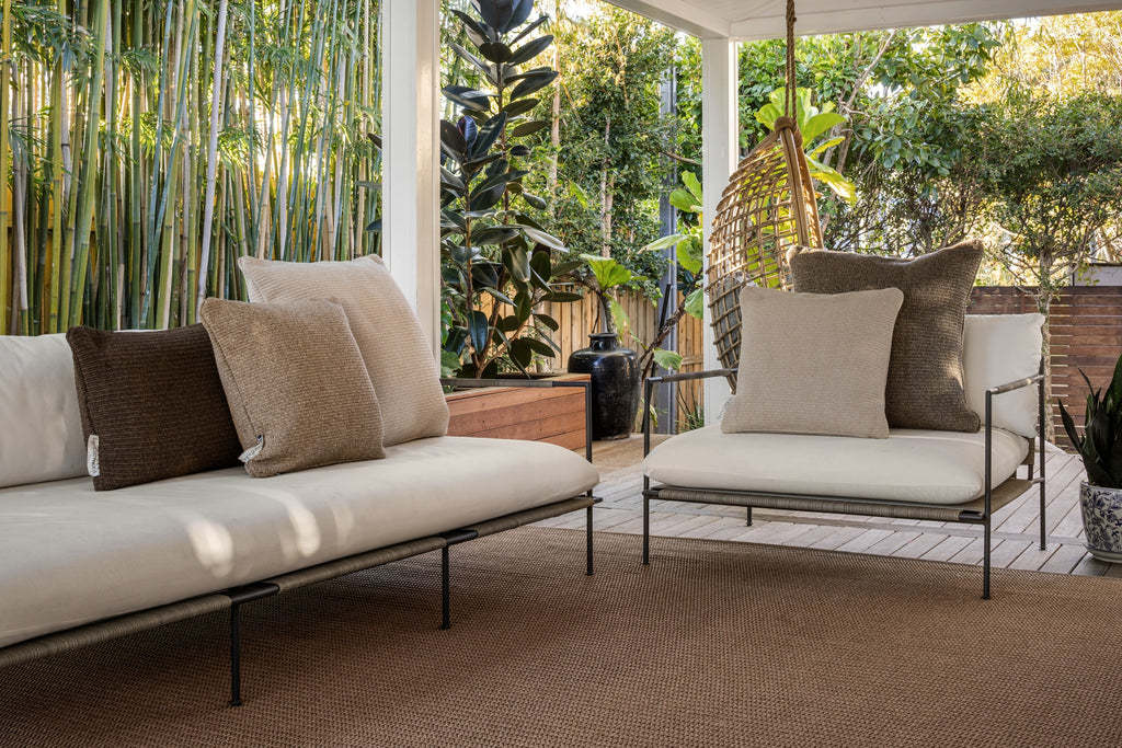 By Carmont Huxley cushion cover in Cameo on an outdoor sofa with brown and tan cushions, beside an armchair