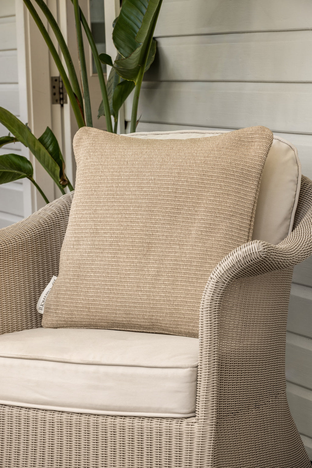 By Carmont Huxley cushion cover in Cameo on a pale wicker armchair against a weatherboard wall