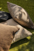 By Carmont Huxley cushion cover in Topaz leaning on a pale cushion on grass, with black and brown cushions