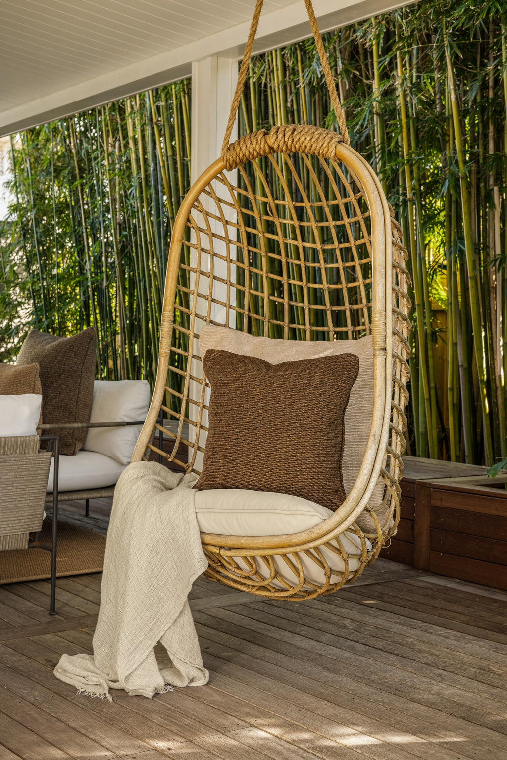 By Carmont Wren cushion cover in Topaz on a hanging chair, angled view with an outdoor sofa behind
