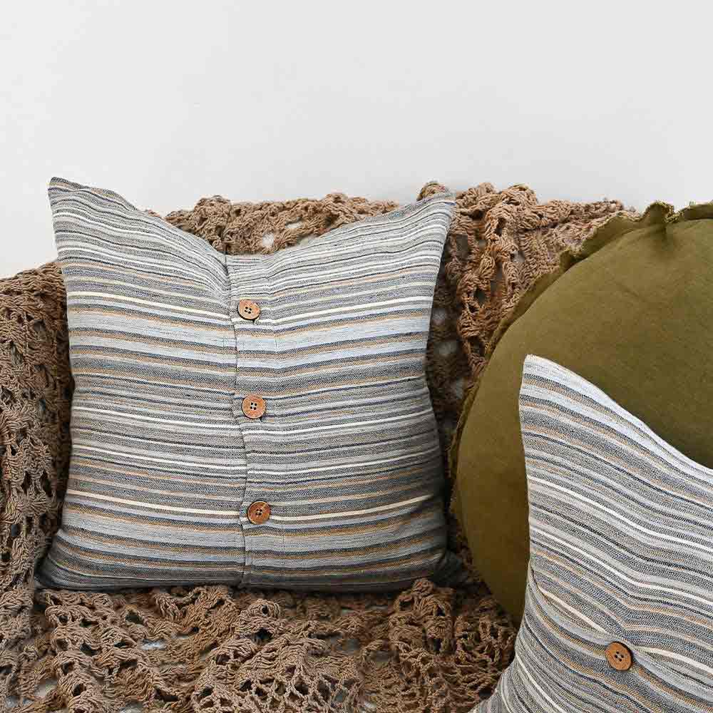 A cushion sitting on a couch with stripes in muted colours of ochre, slate, and red. The front is closed with three wooden feature buttons, and the couch is draped in a fashionable wool throw blanket by Eadie