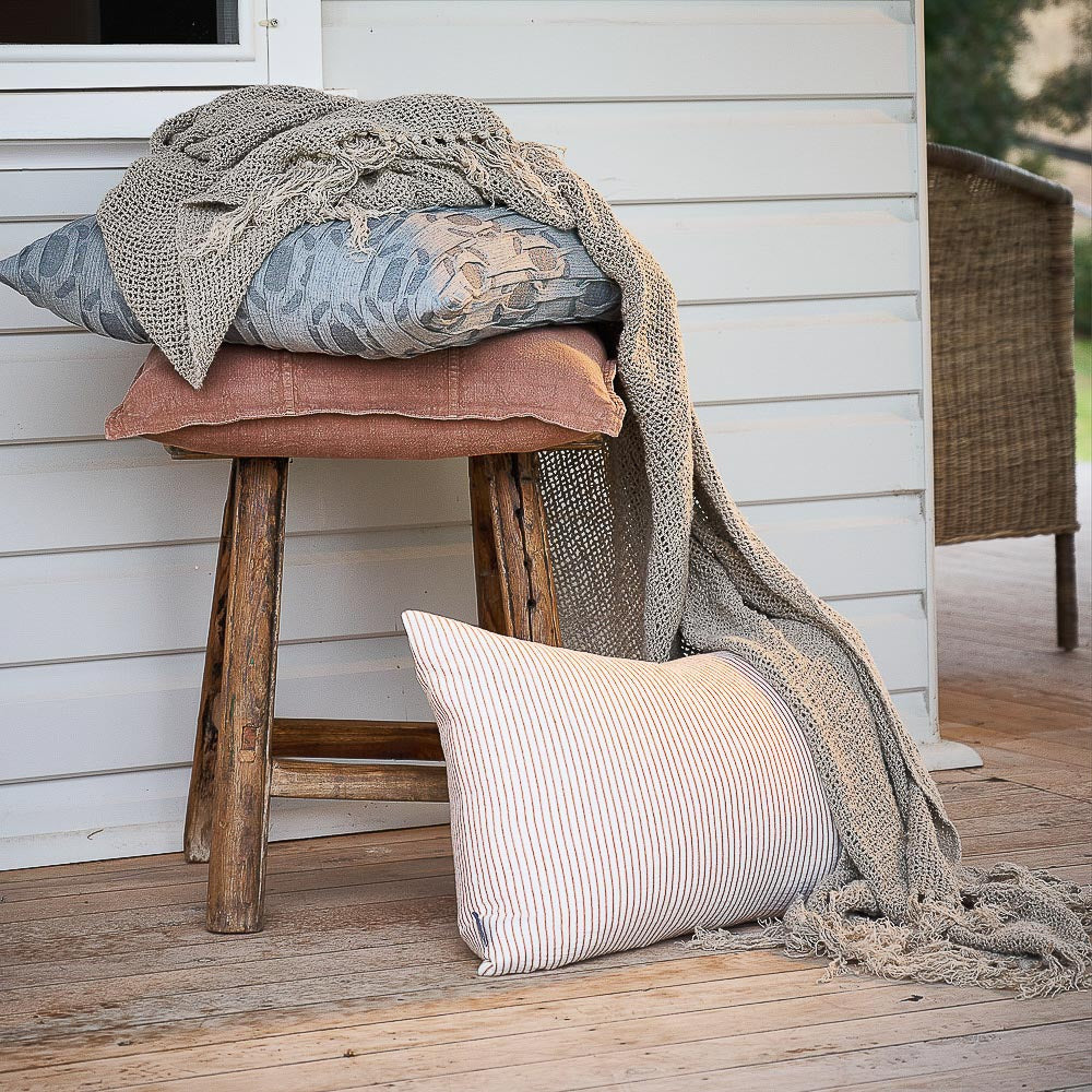 Cushions and a knitted throw on a timber stool on a verandah, with a Luca cushion in Clay.