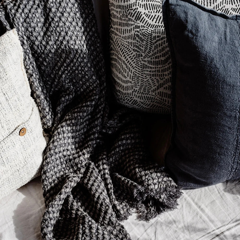 Eadie Lifestyle Luca linen cushion in Midnight on a bed with a woven throw and textured cushions.