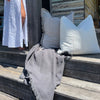 Luca linen cushion in Off White on weathered timber steps beside a fringed grey cushion.
