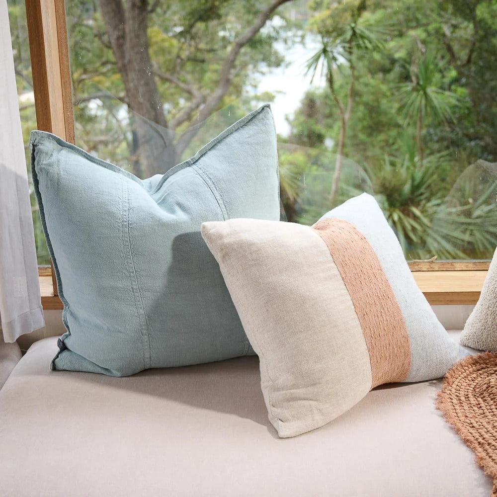 Eadie Lifestyle Luca linen cushion in Sky on a window seat beside a striped cushion.