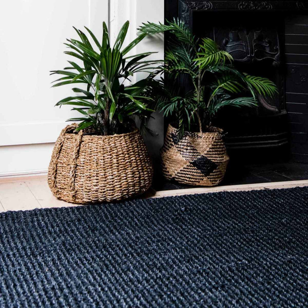 A pair of houseplants atop a large black jute floor rug handwoven in a textured boucle style. 
