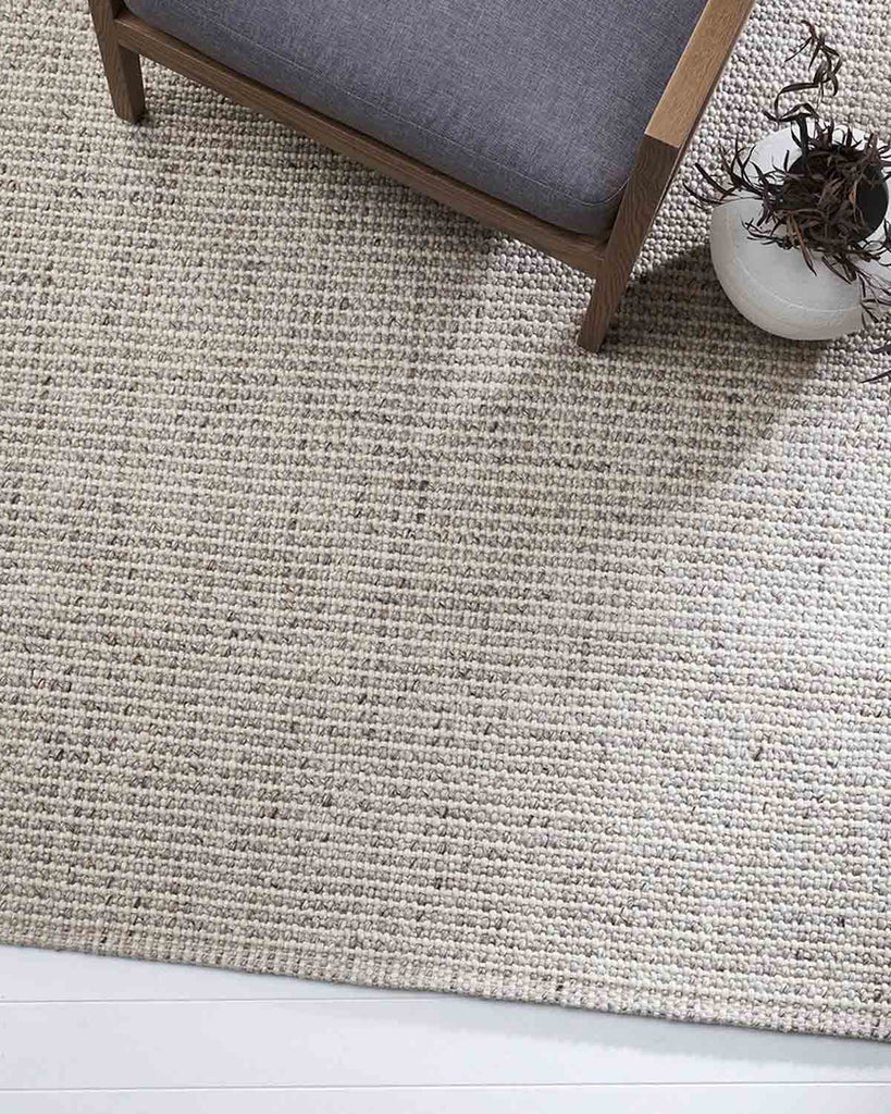 Top-down view of a woven, cool toned neutral Baya NZ rug with a chair. 