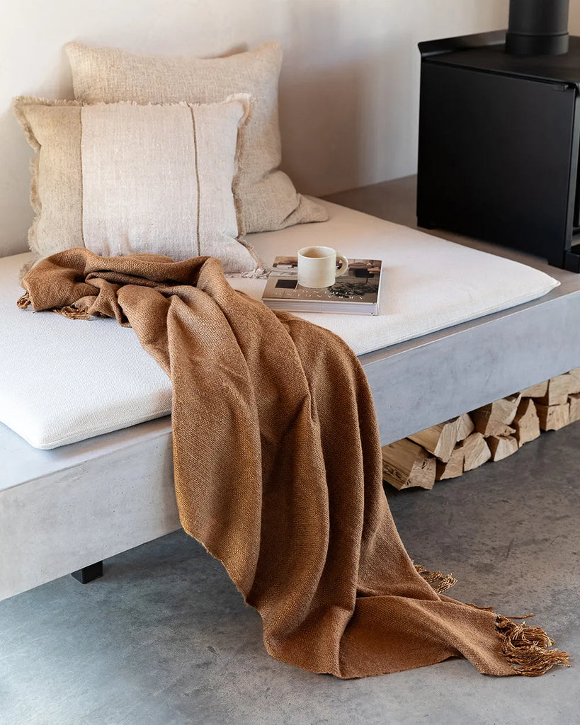 A natural linen cushion with a sand-coloured stripe and fringed edge detail, seen with a brown throw