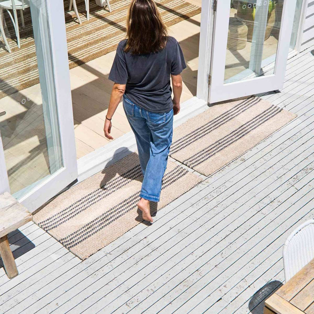 A woman walks into an entryway over two neutral beige coloured doormats, both with striping details in a blue grey tone. 