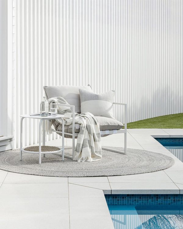 A comfortable looking chair with throws and cushions by Baya NZ, sitting on a grey outdoor rug by a blue swimming pool.