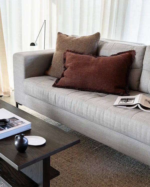 A rich red-brown lumbar cushion with fringe detail, on a couch in a modern living room