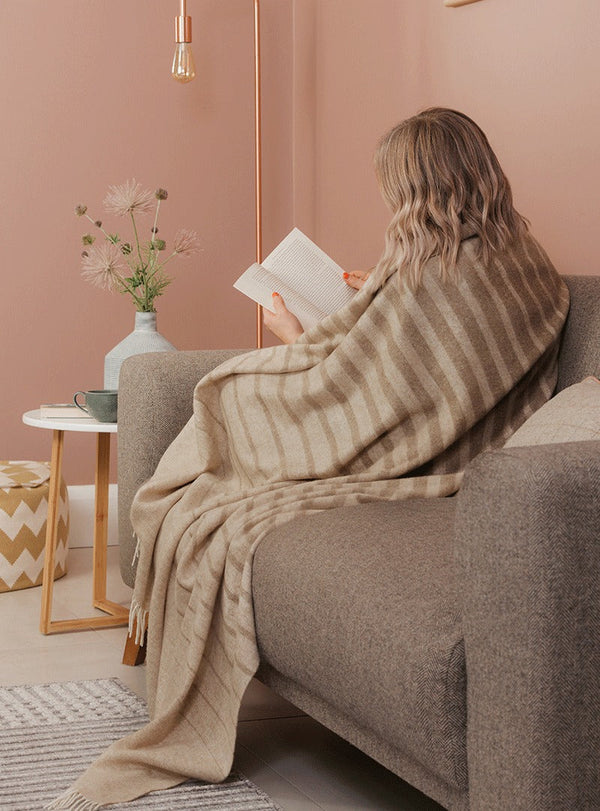 NZ merino wool throw blanket featuring a cream and beige stripe design, seen wrapped around a lady in her home