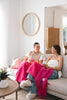 Two women drinking coffee and laughing in a stylish modern living room. There is a bright pink Ruanui Station throw blanket across their knees. 