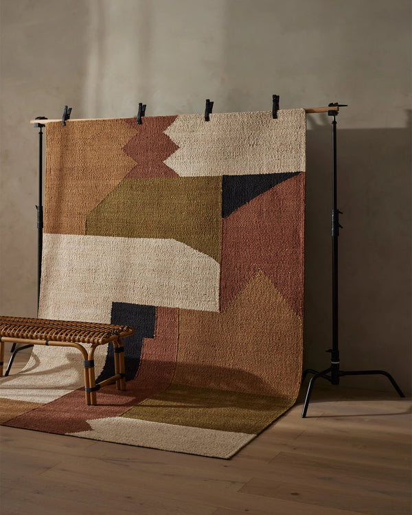 Weave Home Ruben jute rug in Terra hung on a stand in a studio, with a cane bench