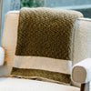 A beautifully textured knit olive green throw blanket with a white striped detail hangs over the back of a stylish modern armchair